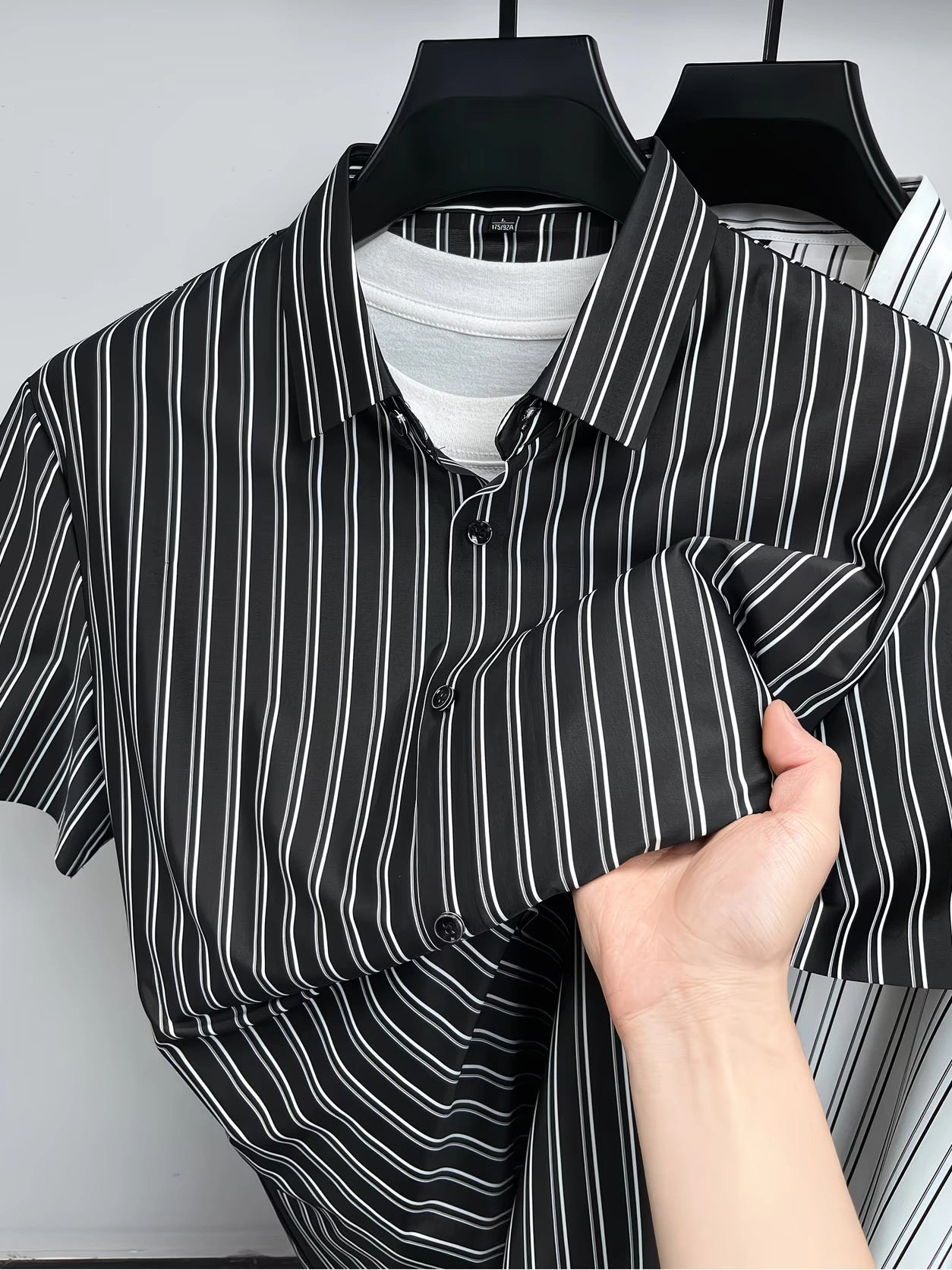 Close hanger view of the GKSEN black white pinstripe men’s shirt showing the collar, button placket, short sleeves, and folded fabric detail.