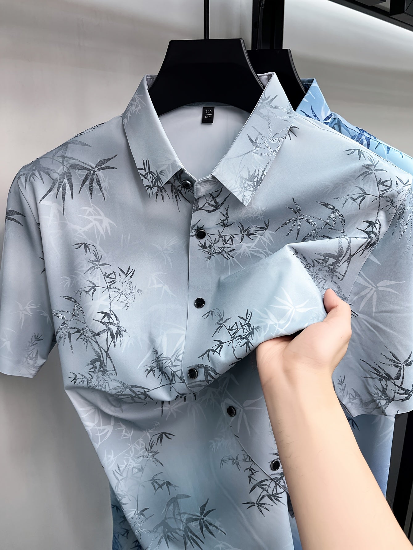 GKSEN silver grey botanical print men’s shirt shown on a hanger with collar, black buttons, folded fabric, and bamboo leaf pattern visible.